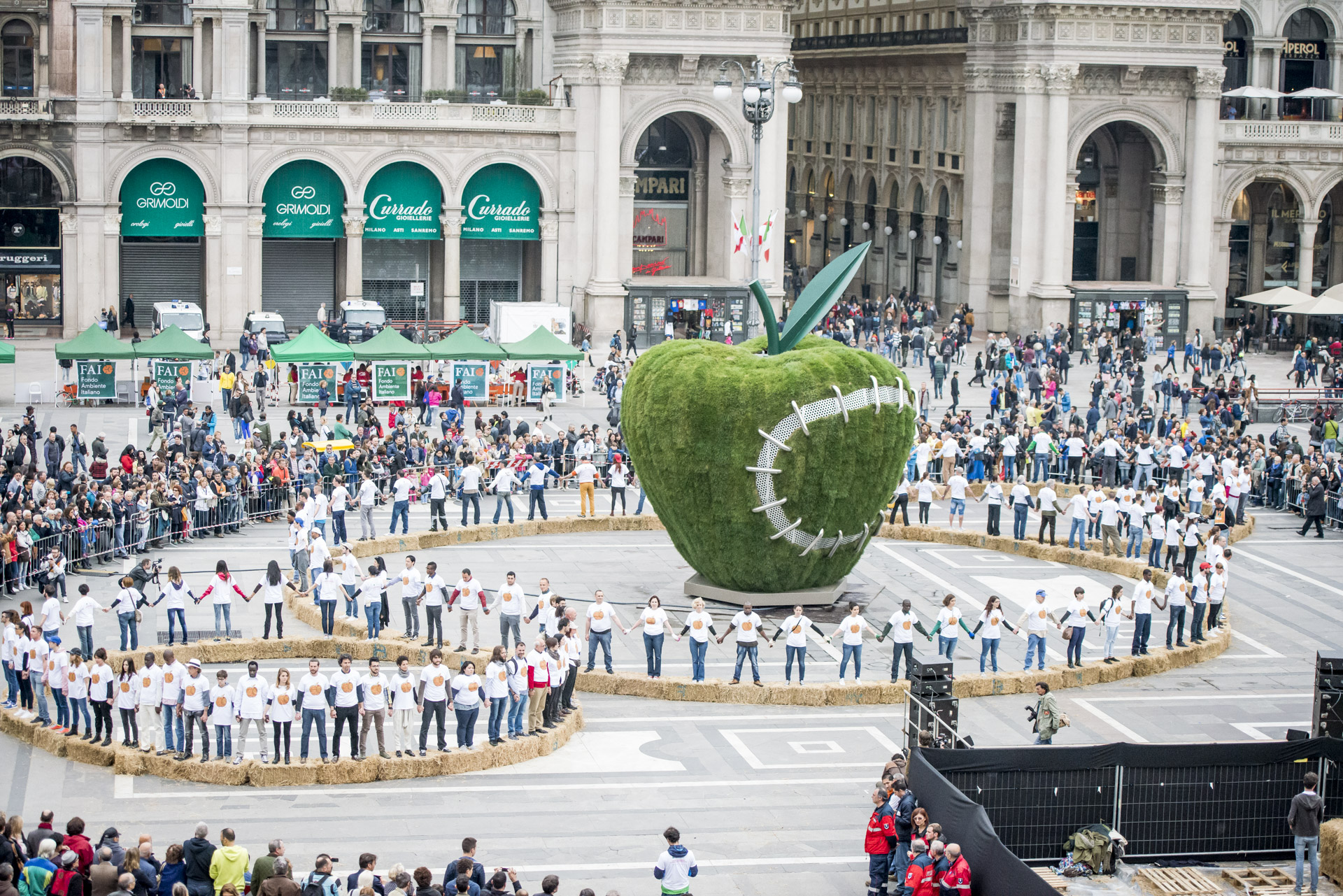 Photo of people standing holding hands forming three circles, Michelangelo Pistoletto Third Paradise symbol, large sculpture of a mended bitten green apple in center, Piazza del Duomo, Milan